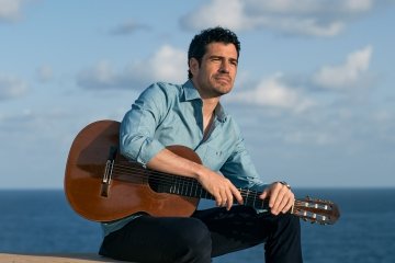 Pablo Villegas sits in front of the ocean, a blue cloudy sky behind him. His guitar is propped on his lap,