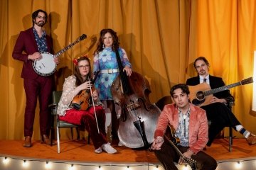 5 musicians in various poses on an orange stage with an orange stage curtain. Missy raines stands in the middle with her bass