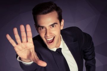 Michael Carbonaro holds his hand up to the camera and smiles widely