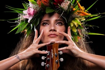 Taimane, wearing a floral headdress, poses with her hands splayed across her face and a Ukulele in front of her