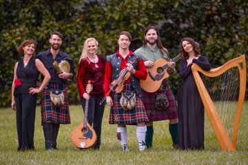 6 celtic band members pose on the grass, standing side by side and holding various instruments including a harp and guitar.
