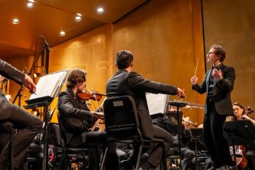 Teddy Abrams stands to the right of frame, hands in the air as he conducts the orchestra in front of him. You can see violinists and bassists playing