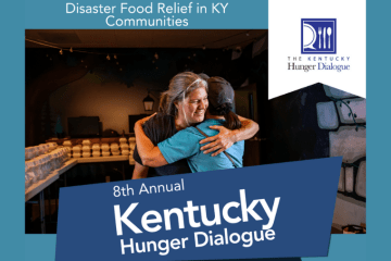Two people embracing and smiling at a food distribution center for disaster relief. In the background are tables with food boxes.