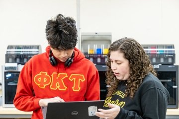 Student talking with professor holding laptop. Standing in front of three 3rd printers