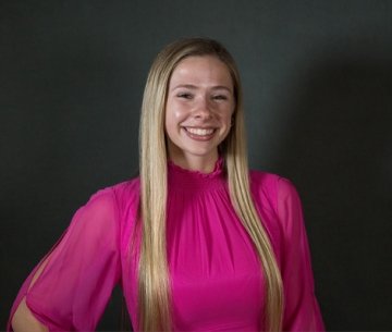 Student wearing pink top standing in front of dark background smiling at camera