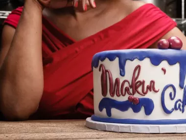 A woman sitting behind a cake which says, in frosting "Makin' Cake"