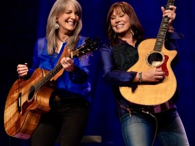 Kathy Mattea and Suzy Bogguss each playing an acoustic guitar and smiling