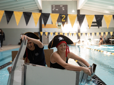 Two students in pirate hats paddle a cardboard box in a large swimming pool.