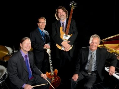 The Brubeck Brothers Quartet. One man sits by a drum, two stand with guitars, and one sits by a piano.