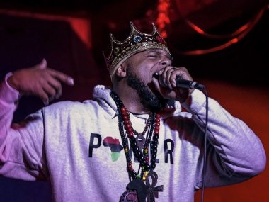 Devine Carama rapping into a microphone. He has necklaces and a crown on.