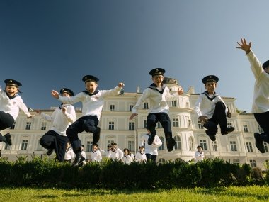 Members of the Vienna Boys Choir jumping over a bush, smiling and throwing their hands in the air