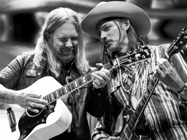 A black and white image of two men playing guitar. The man on the right is wearing a cowboy hat and is leaning over to the man on the left and speaking.