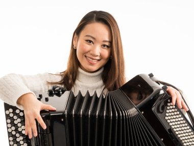 A brown haired woman sits in a white turtle neck holding an accordion in her lap with a white background.