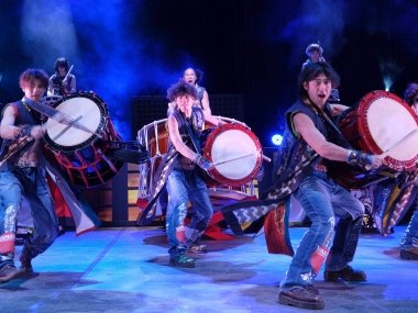 Among the eight drummers on stage, three are centrally located on the stage in front of the camera. These drummers are all dressed in blue jeans, black mid-calf length jackets, and have large red drums under their left arms that they are drumming on.