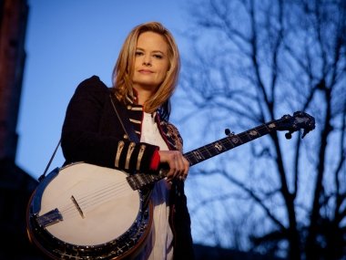 A blond woman stands in front of a blue sky wearing a black jacket and white shirt holding a banjo stapped over her right shoulder.