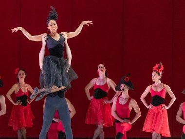 Seven dancers dressed in French-styled red dresses and hats stand in front of a red curtain. In front of those dancers, there is a male and female dance pair dressed in black. The woamn is raised above the man with her arms spread outwards and feet kicked up behind her knees.