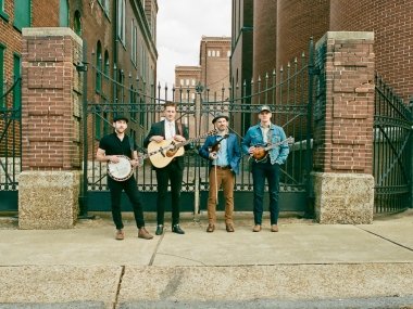 Four men stand in front of copper green gates in a street of brick buildings. The men (starting from the leftmost) hold a banjo, guitar, violin, and electric guitar under their right arms.