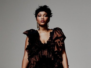 Luedji Luna (a woman with short, black curly hair) in a dark red and black dress in front of a gray background.