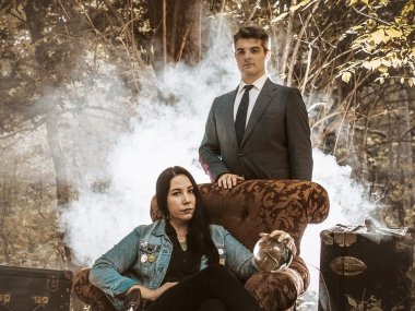 A woman with long, black hair sitting in a high back chair holding a crystal ball. A man in a grey suit is standing behind the chair. The background is primarily trees and branches, but is partially covered by the fog behind the man.