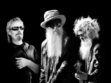 A balck and white image of three men. The left-most man has a mullet and sunglasses, the middlemost man has a long beard, hat and sunglasses, and the rightmost man has a mid-length beard, sunglasses, and is playing the guitar.