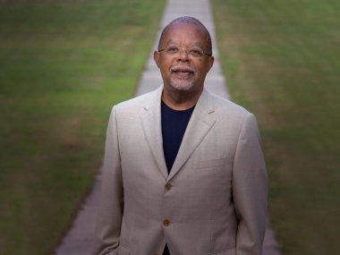 Henry Louis Gates Jr. is standing in front of a blurred sidewalk in grass. He is wearing a grey suit and black shirt.