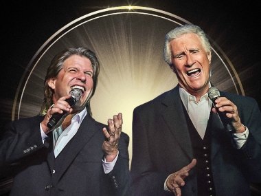Two men in grey suits in front of a black background with a golden halo and light radiating from the center position of the image. Both men are holding microphones, and the photo is taken while they are singing.