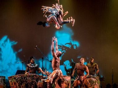 Acrobat performers on stage. One flips in the air as three stand on the floor. Two are behind the performers playing the drums
