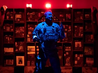 A man in a military uniform stands bathed in blue light, in front of a bookcase full of memorabilia which is bathed in red light.