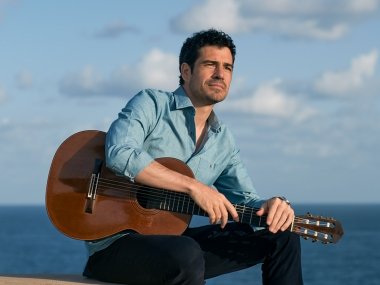 Pablo Villegas sits in front of the ocean, a blue cloudy sky behind him. His guitar is propped on his lap,
