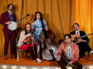5 musicians in various poses on an orange stage with an orange stage curtain. Missy raines stands in the middle with her bass