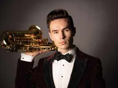 Valentin, dressed in a suit, stands in front of a dark background holding up a saxophone leaning against his shoulder