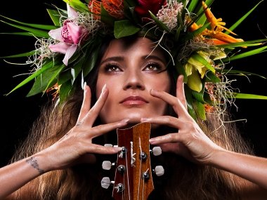 Taimane, wearing a floral headdress, poses with her hands splayed across her face and a Ukulele in front of her