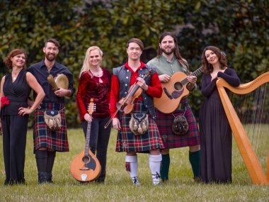 6 celtic band members pose on the grass, standing side by side and holding various instruments including a harp and guitar.