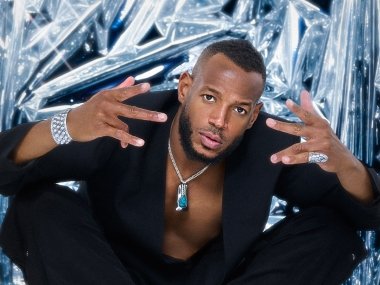Marlon Wayans sits cross-legged on a floor with a shiny metal backdrop behind him