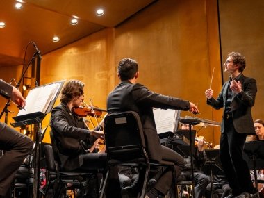 Teddy Abrams stands to the right of frame, hands in the air as he conducts the orchestra in front of him. You can see violinists and bassists playing