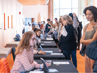 Students sitting at tables presenting to attendees walking past each table in an open house style event.