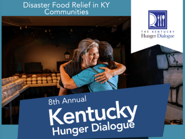 Two people embracing and smiling at a food distribution center for disaster relief. In the background are tables with food boxes.