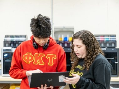 Student talking with professor holding laptop. Standing in front of three 3rd printers