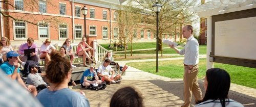 Students listening to a professor in an outdoor classroom with white board behind the professor