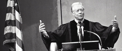 Former President Jimmy Carter spreads his arms wide as he speaks behind a podium.