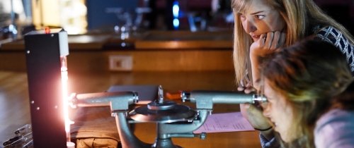 One female student watches as another looks through a scientific tool.