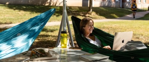 A student uses a laptop computer while lying in a hammock suspended in the shade. A water bottle and other items are on a table nearby.
