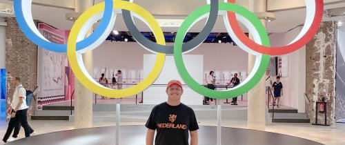 A man wearing a t-shirt and baseball cap poses in front of the Olympic Rings.