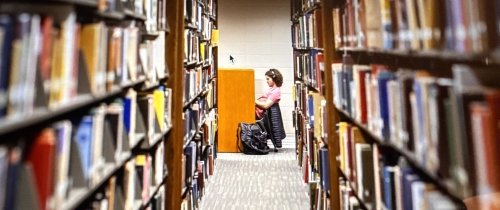 inside Grace Doherty Library with stacks of books on each side of row and student studying at end of row