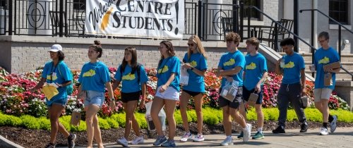 Nine college students wearing matching blue t-shirts pass a banner hanging from a downtown building that states &quot;Welcome Centre Students.&quot;