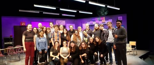 A group of student actors, along with faculty members post for a photo on stage in front of a set designed to look like a high school classroom.
