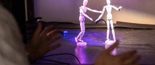 Two posable miniature figures are seen on a model stage as a man's hands gesture in the foreground.