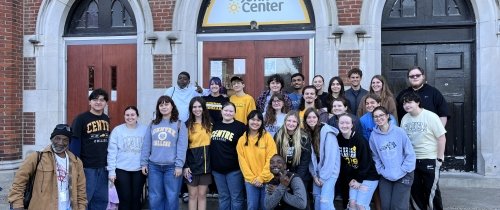 A group of more than 20 young men and women, many wearing Centre College clothing, pose for a photo on the step of a brick building, upon which a sign reads "St. John Center."