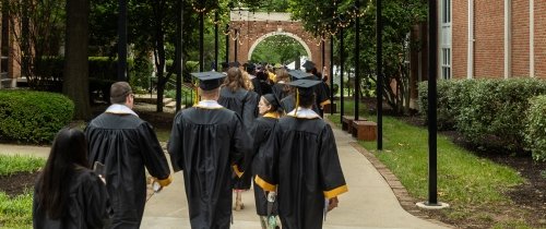 Centre College students wearing commencement robes and caps make their way through a brick archway on campus.