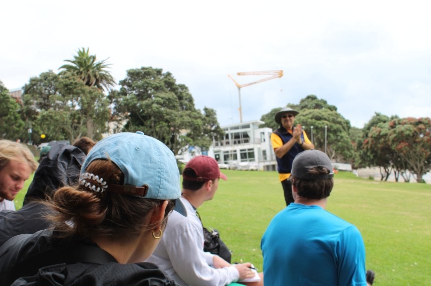 A professor speaks with a group os students seated on a green lawn.